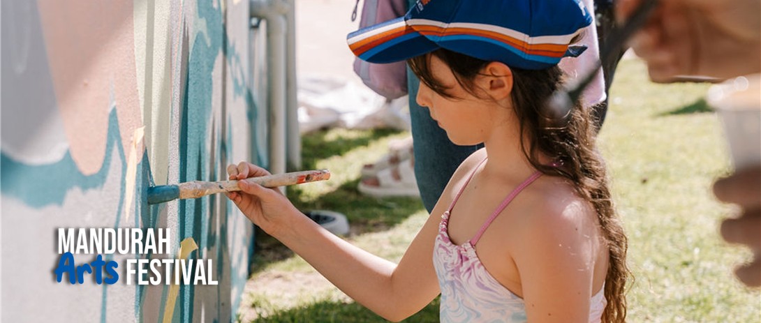 A young girl kneels on the ground outdoors in front of an exterior wall, holding a paintbrush and filling in a space on a mural with blue paint